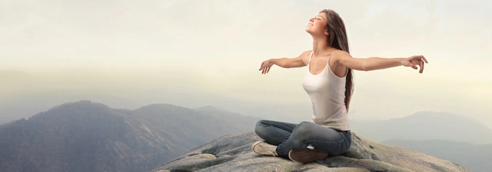 A person seated on a mountaintop with arms open, embracing calm and expansive natural views.